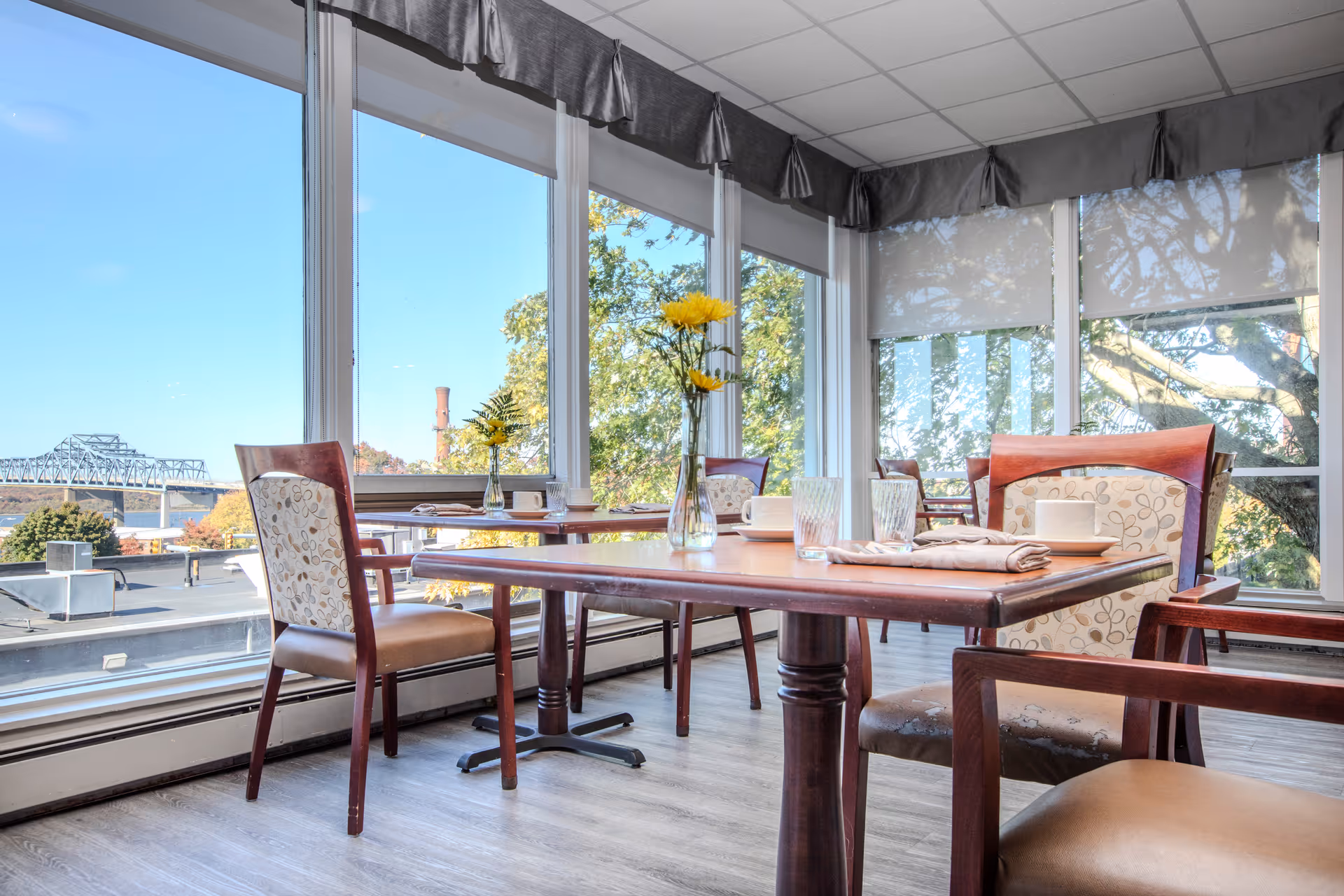 A bright dining area with wooden tables and cushioned chairs featuring floral patterns. The room has large windows offering a view of trees, a bridge, and a clear blue sky. Each table is set with glasses, plates, napkins, and a vase with yellow flowers.