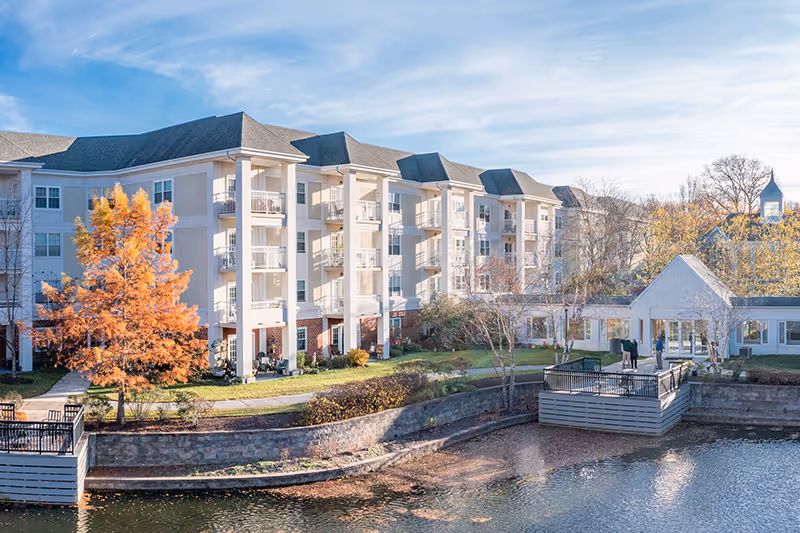 Exterior view of a multi-story senior living facility named The Evergreens, featuring balconies, a landscaped garden with autumn-colored trees, and a waterfront area with a small dock under a blue sky.