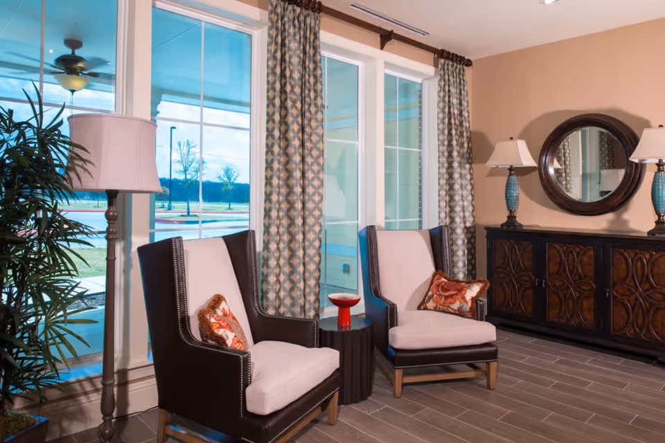 A bright seating area with two upholstered armchairs and a small side table in front of large windows, flanked by lamps, a decorative cabinet, and a round mirror.