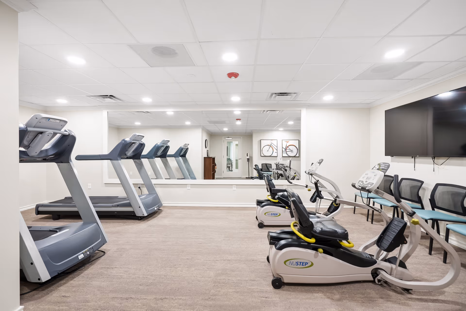 A fitness room with several exercise machines including treadmills and recumbent bikes. There is a large mirror on the back wall reflecting the equipment and a door. On the right side, there is a large flat-screen TV mounted on the wall above a row of chairs. The room has a beige carpet and white ceiling with recessed lighting.