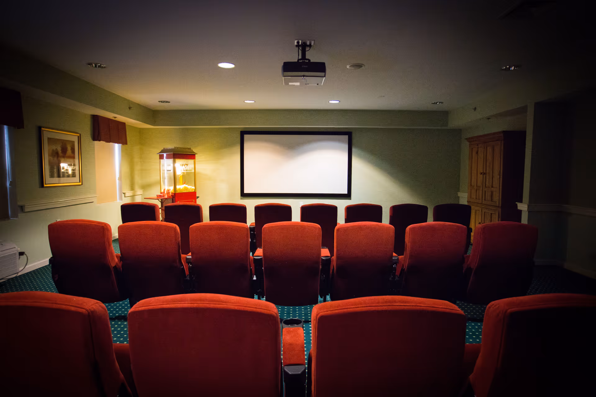A small movie theater room with rows of red cushioned chairs facing a blank projection screen. There is a popcorn machine on the left side near the wall, and a wooden cabinet on the right side. The room has green carpet with a dotted pattern and light green walls.