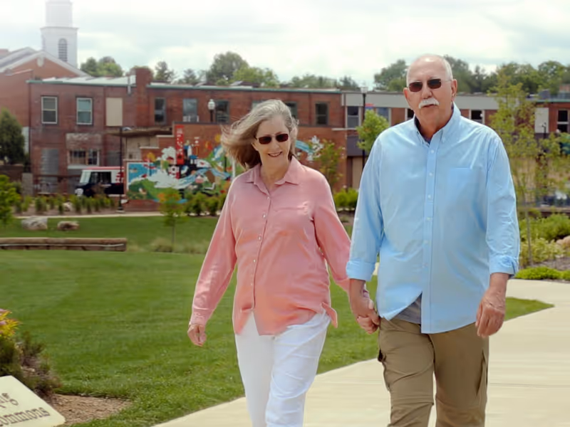 An elderly couple holding hands and walking on a paved pathway in a green outdoor area with buildings and a mural in the background. The woman is wearing a pink shirt and white pants, and the man is wearing a light blue shirt and khaki pants. Both are wearing sunglasses.