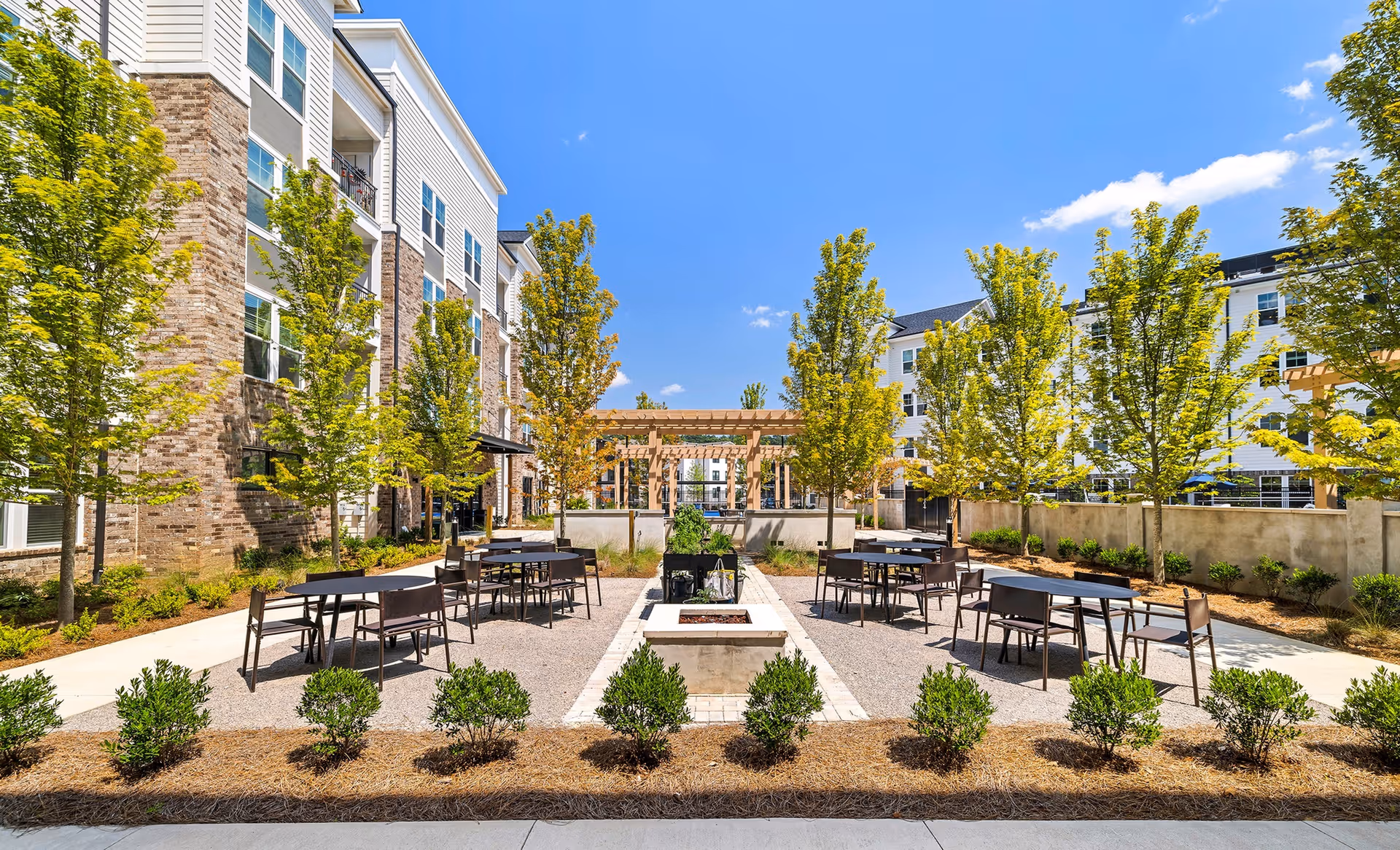 Outdoor courtyard area at Overture Tributary featuring multiple black tables and chairs arranged around a central fire pit, surrounded by young trees and shrubs, with multi-story residential buildings in the background under a clear blue sky.