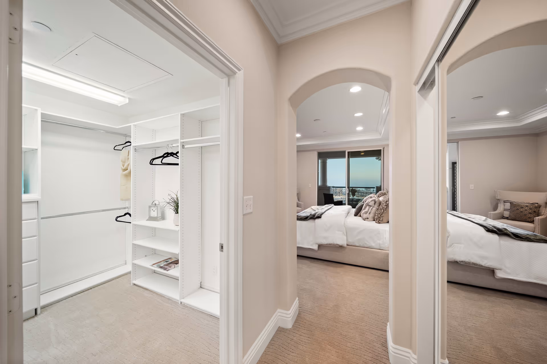 View of a spacious walk-in closet with white shelves and hanging rods, adjacent to a bedroom with a large bed, decorative pillows, an armchair, and a sliding glass door leading to a balcony with an ocean view.