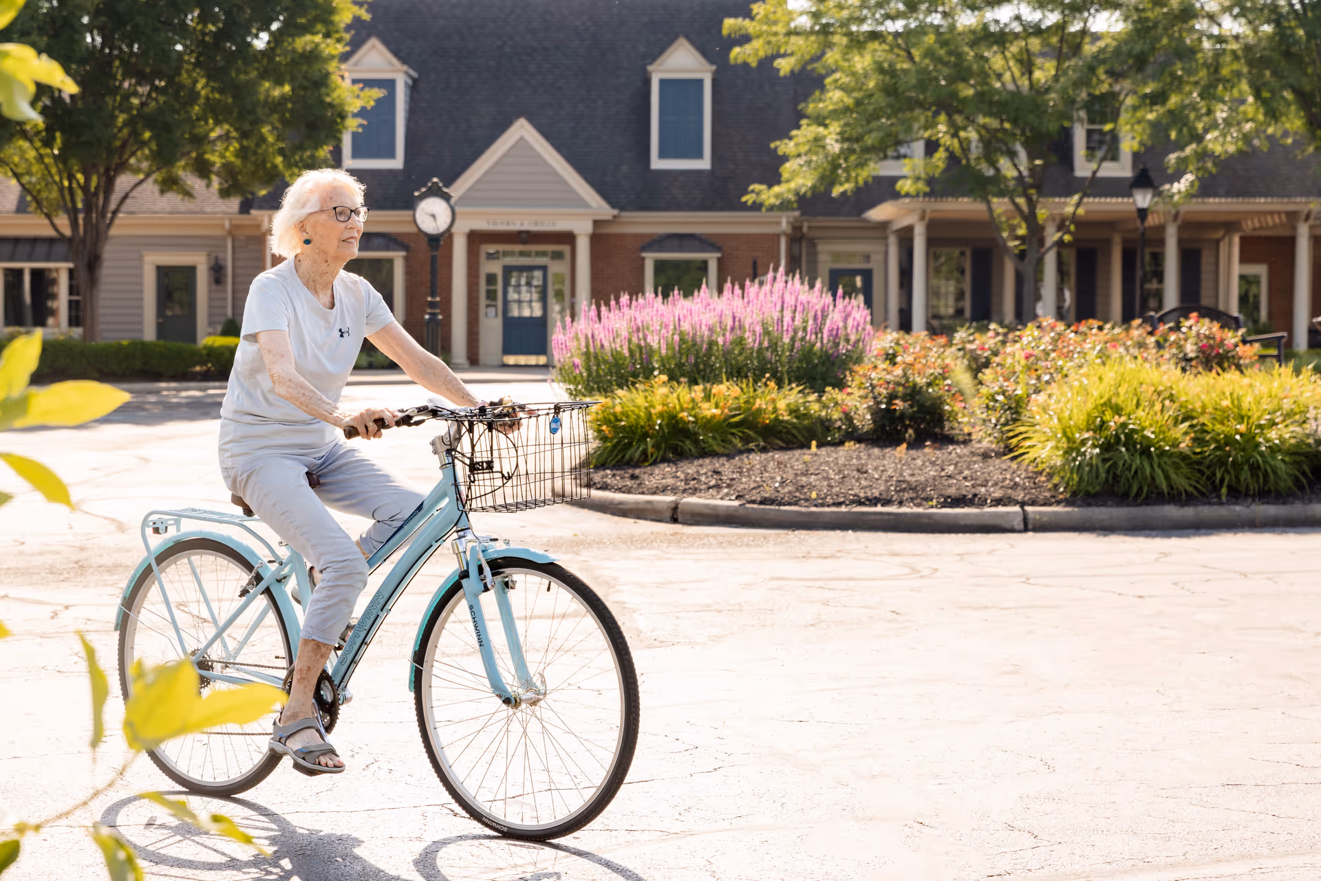 An elderly woman with white hair and glasses rides a light blue bicycle in a sunny outdoor area with trees, flowering bushes, and a building with a clock in the background.
