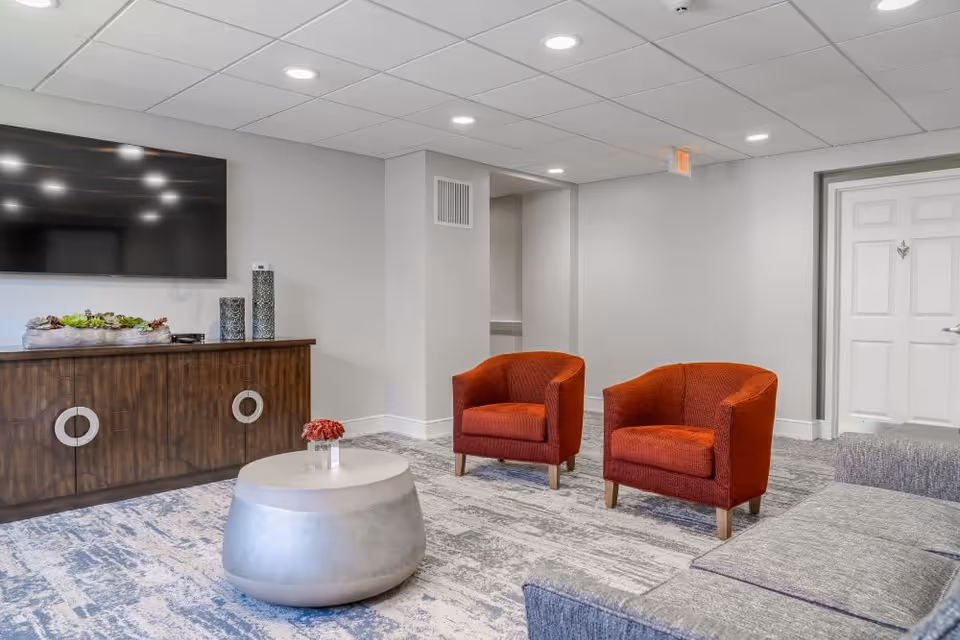 A modern living room area with two red armchairs, a gray sofa, a round silver coffee table with a small flower vase, a wooden cabinet with decorative items, and a large flat-screen TV mounted on the wall. The room has a light gray carpet and white walls with recessed ceiling lights.