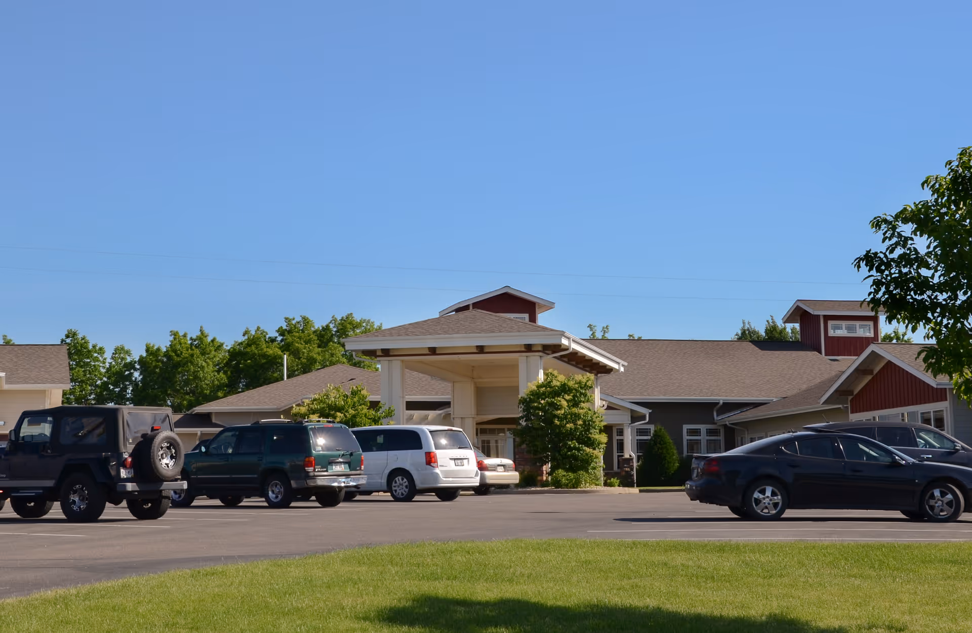 Exterior view of Spring Valley Senior Living and Health Care Campus building with a covered entrance and several parked cars in the parking lot under a clear blue sky.