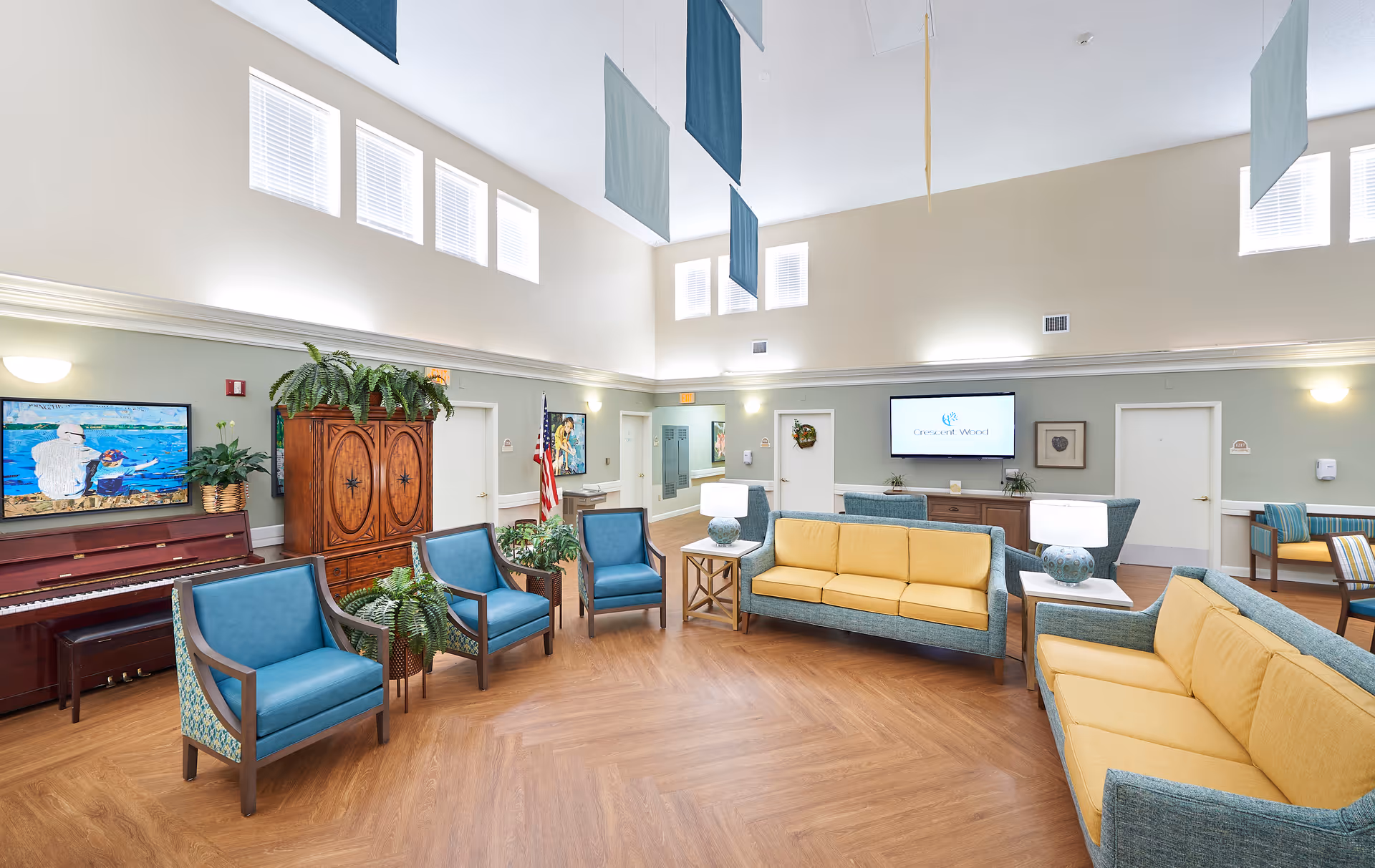 Bright communal lounge with yellow sofas, blue armchairs, a piano, TV, plants, and high ceilings.