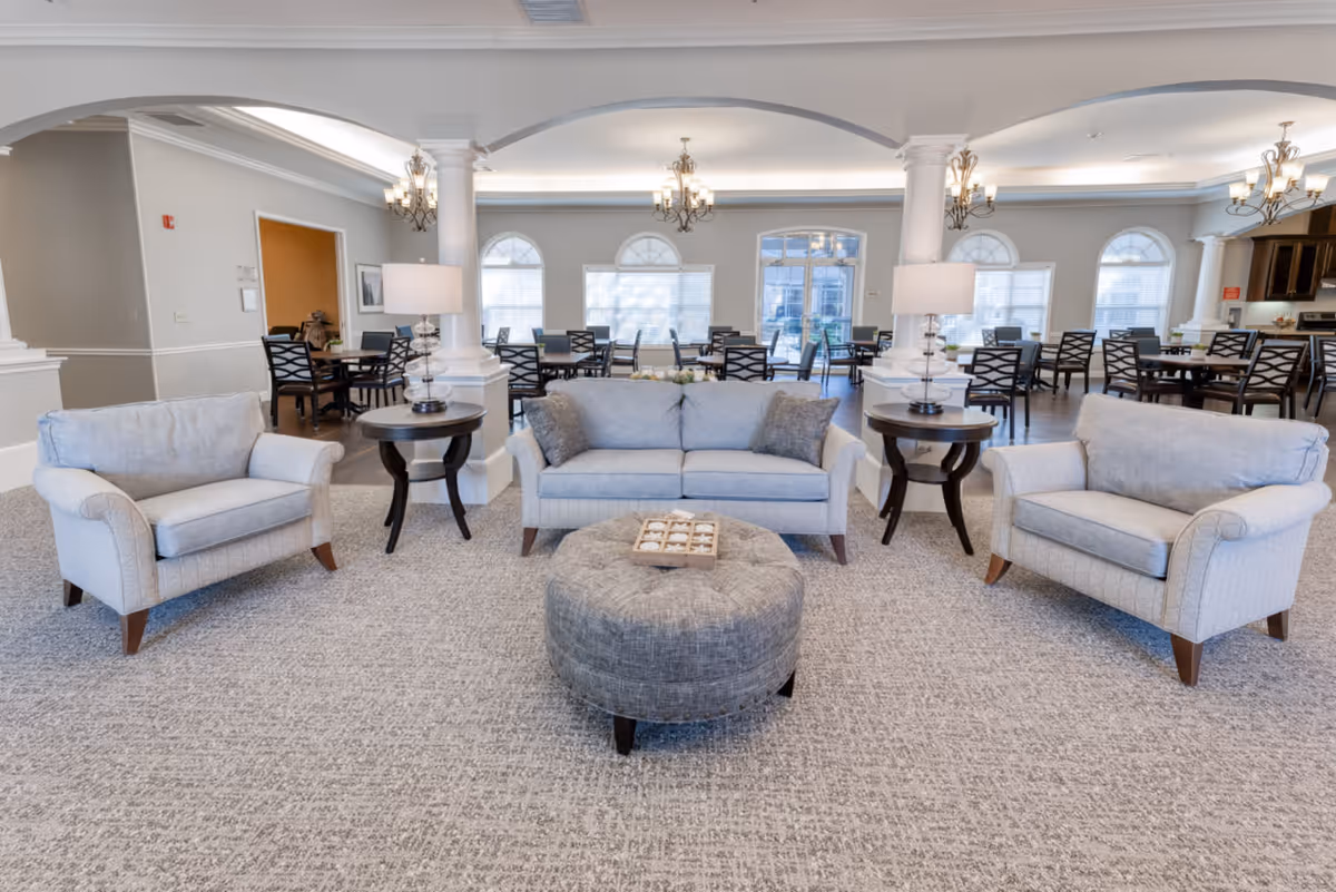 A spacious senior living facility common area with light gray upholstered seating including a sofa and two armchairs arranged around a round gray ottoman with a tic-tac-toe game on top. Two dark wood side tables with lamps flank the sofa. In the background, there are multiple dining tables and chairs, large windows with arched tops, white columns, and chandeliers hanging from the ceiling.