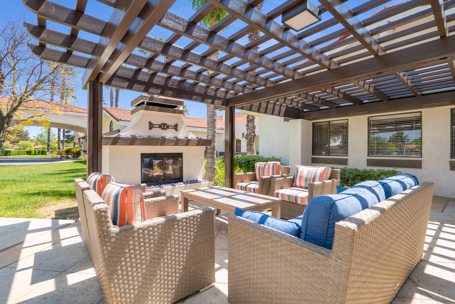 Outdoor seating area with wicker furniture including a sofa and chairs with colorful cushions arranged around a rectangular coffee table under a wooden pergola. A fireplace is built into a stucco wall nearby, and the area is surrounded by green grass and trees with a building in the background.