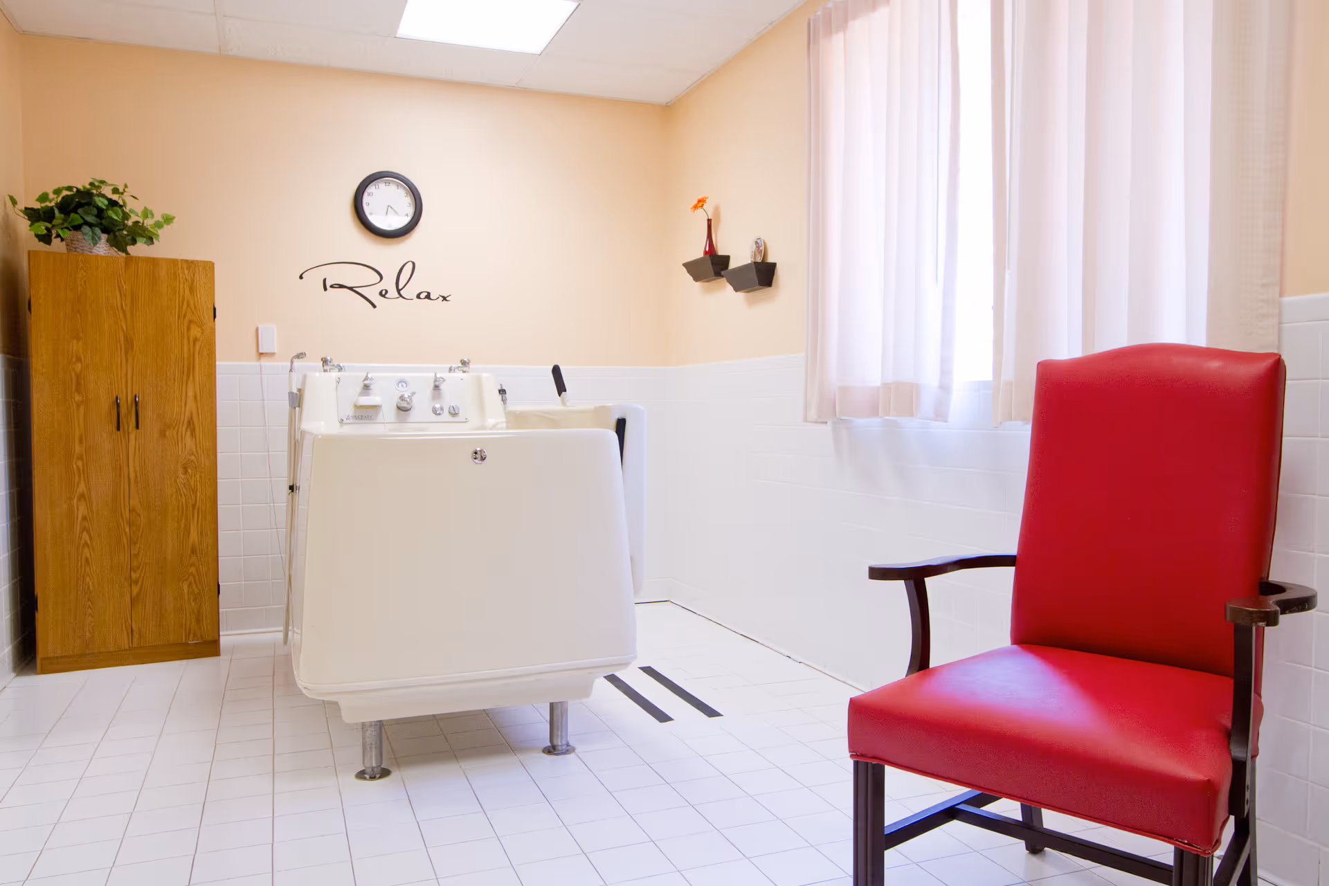 A bright room with white tiled floor and walls halfway up, featuring a large white bathing tub in the center, a wooden cabinet with a green plant on top to the left, a red cushioned chair with wooden arms to the right, and a window with light curtains. On the wall above the tub is a clock and the word 'Relax' written in cursive, with two small shelves holding decorative items.