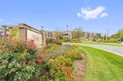 Outdoor view of The Summit senior living facility entrance sign surrounded by landscaped flower beds with red and yellow flowers, green grass, and a clear blue sky with a few clouds. The multi-story building is visible in the background.