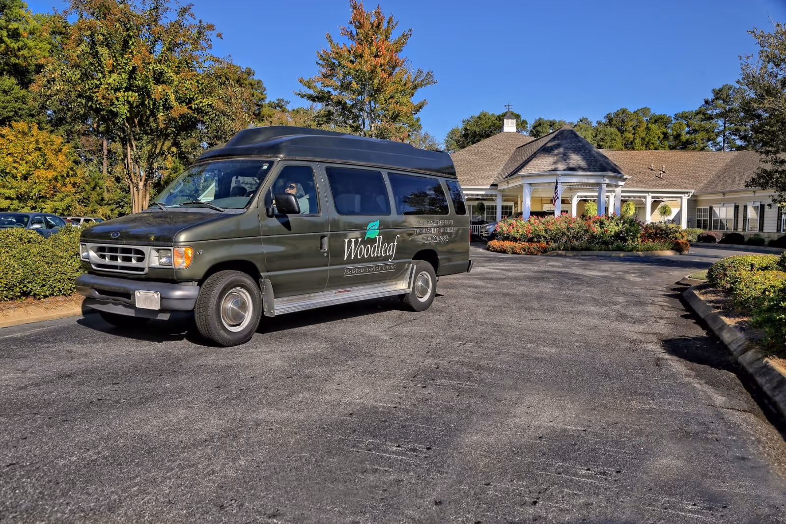 A Woodleaf Senior Care shuttle van parked in front of the single-story senior living facility entrance.