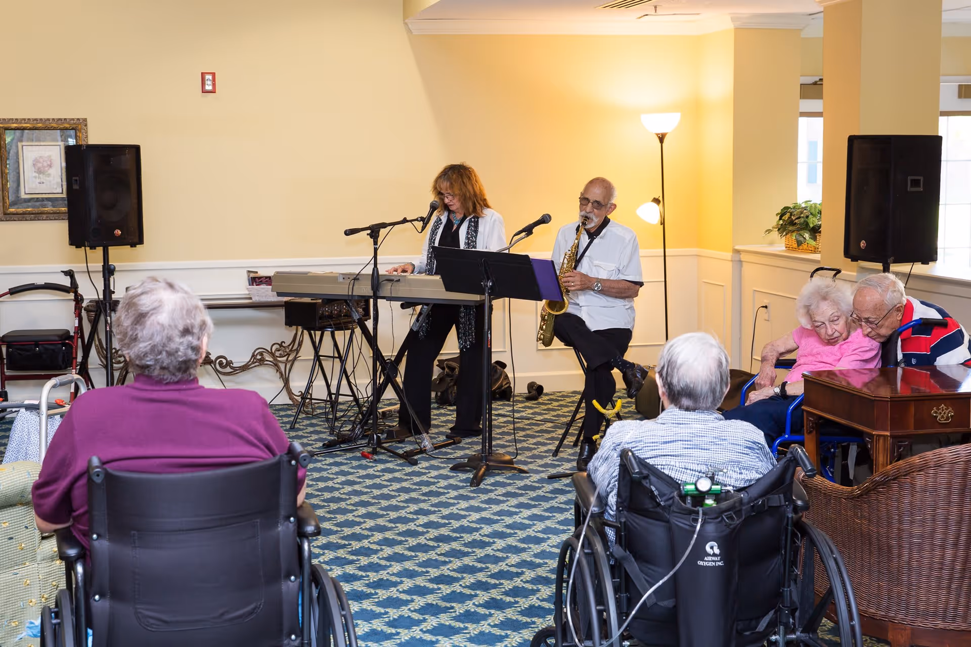 A small group of elderly people, some in wheelchairs, watch a live musical performance in a common room. A woman plays a keyboard and sings into a microphone, while a man plays a saxophone. The room has patterned carpet, yellow walls, and speakers set up for the performance.