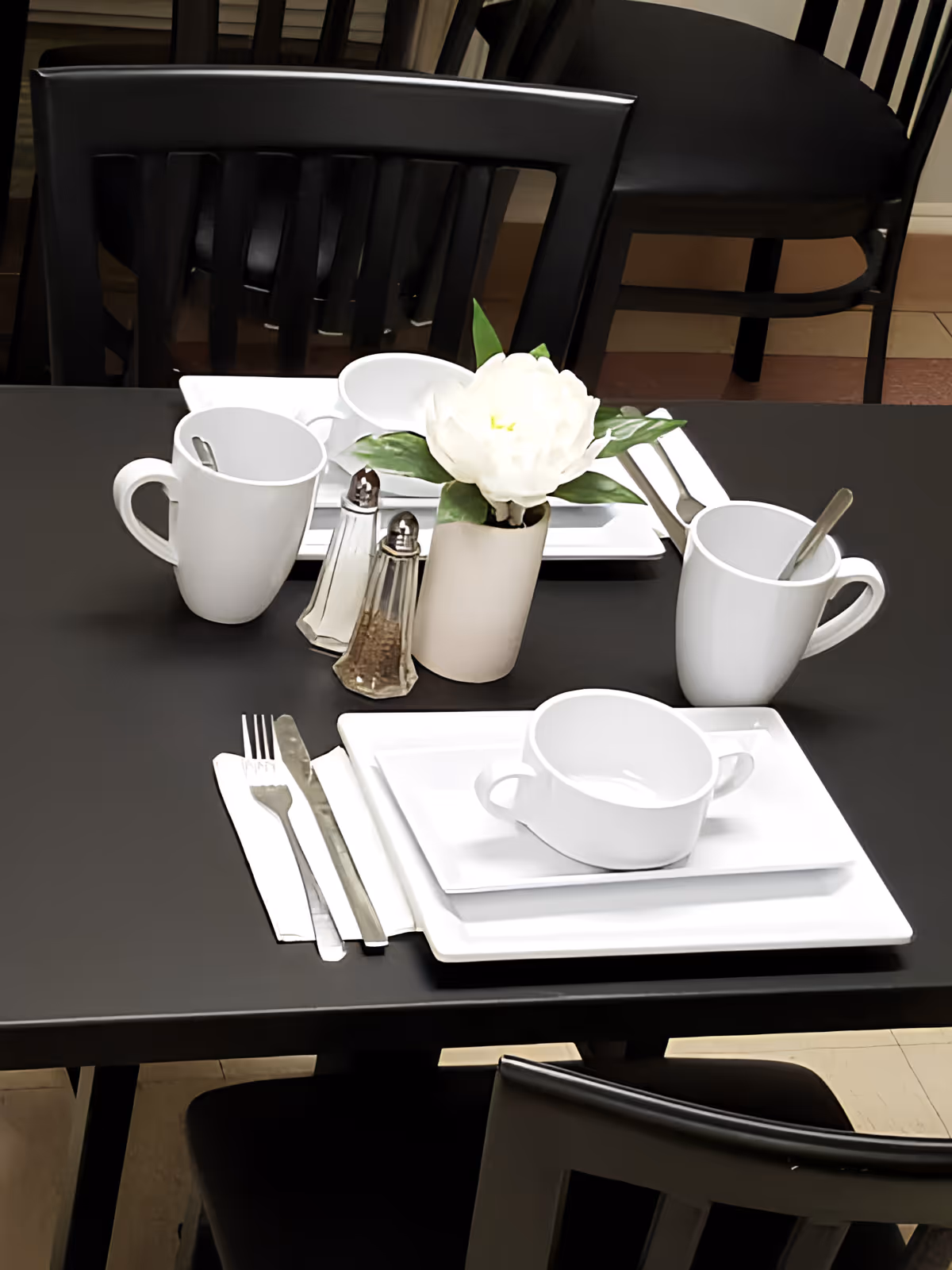 A dining table set with white square plates, a white soup bowl, two white mugs with spoons, silverware on white napkins, a salt and pepper shaker, and a small white vase with a white flower and green leaves. Black chairs surround the table.