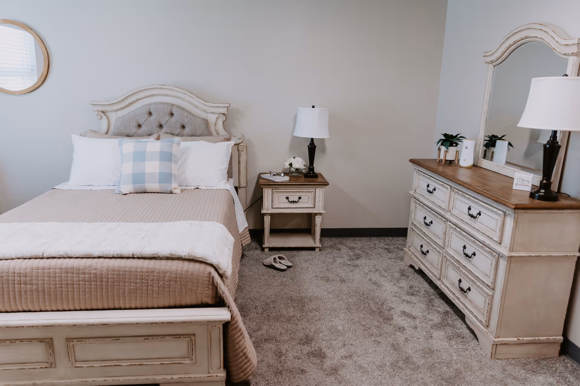 A cozy bedroom with a beige tufted headboard bed, beige bedding, and a blue and white checkered pillow. There is a nightstand with a lamp and decorative items next to the bed. Across from the bed is a matching dresser with a large mirror, two lamps, and small plants on top. The room has carpeted flooring and neutral-colored walls.
