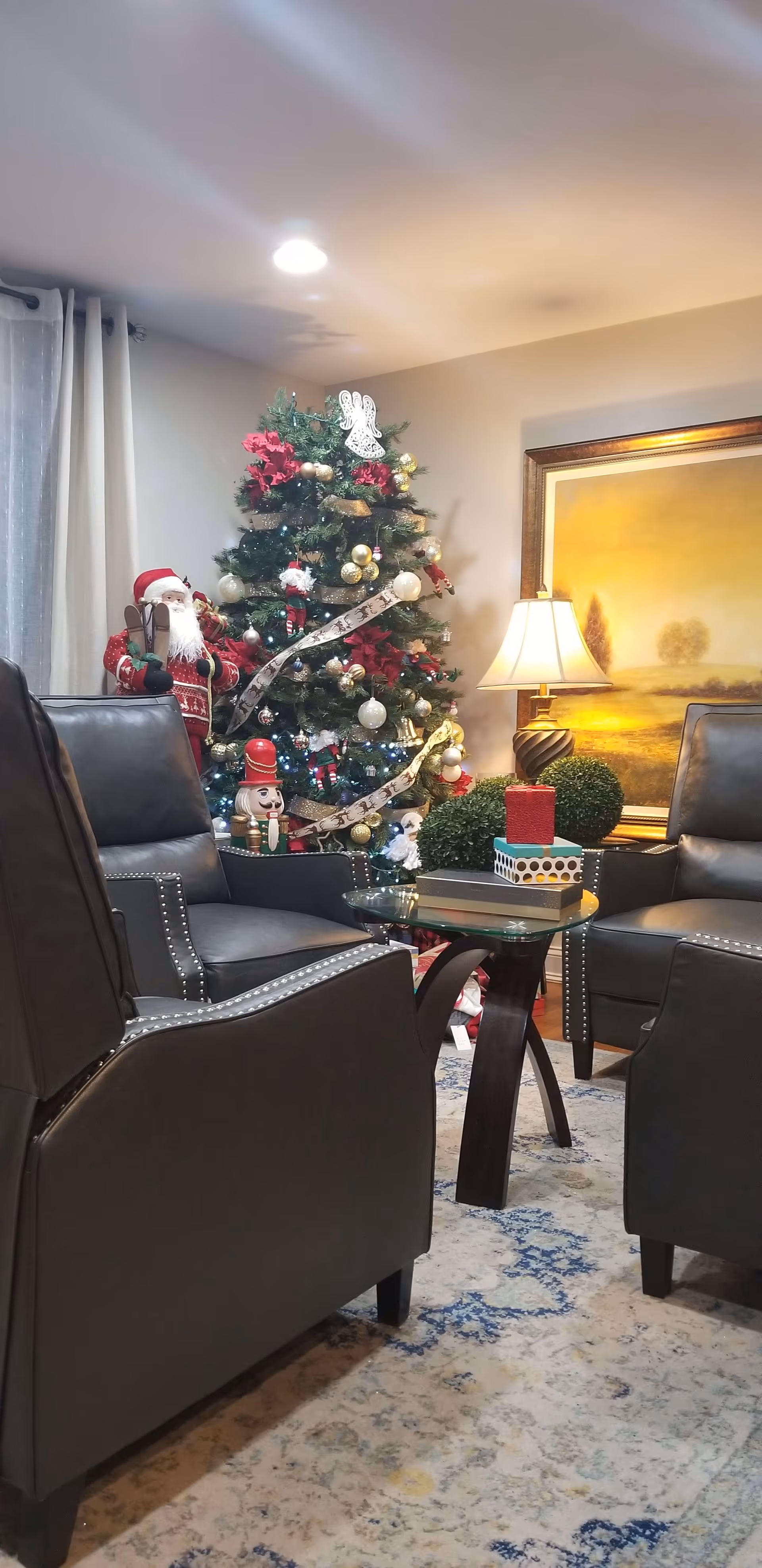 A cozy living room decorated for Christmas with a decorated Christmas tree featuring ornaments, ribbons, and a white angel topper. There are four dark leather armchairs arranged around a glass-top coffee table with wrapped gift boxes on it. A table lamp and a framed landscape painting are visible in the background.
