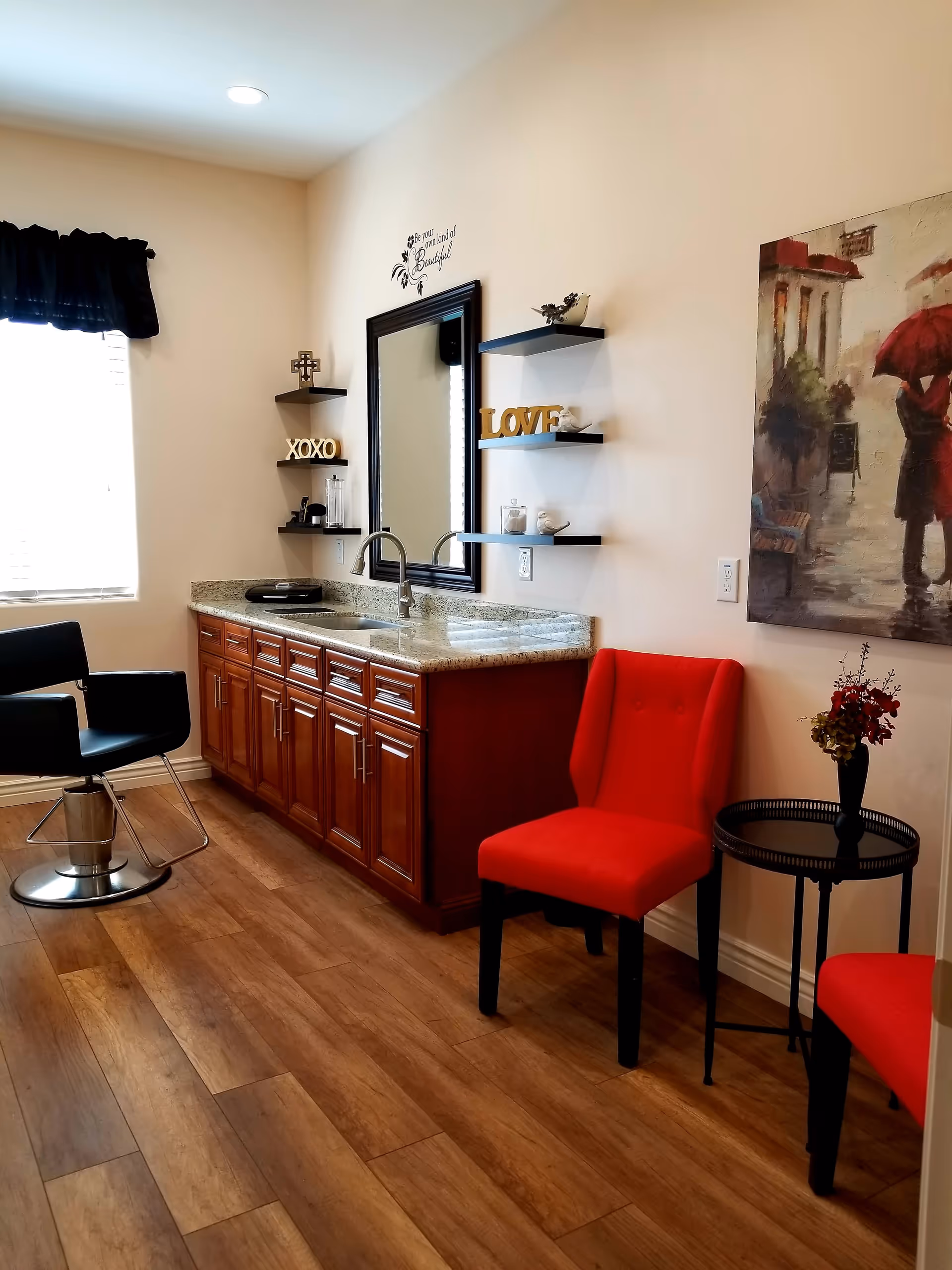 A cozy interior room with wooden flooring featuring a granite countertop with a sink and wooden cabinets underneath. Above the countertop is a large mirror flanked by three black floating shelves holding decorative items including the words 'XOXO' and 'LOVE'. To the right, there are two red upholstered chairs with a small round black table between them holding a vase with flowers. A painting of a person holding a red umbrella is hung on the wall. A black salon-style chair is positioned near a window with a black valance.