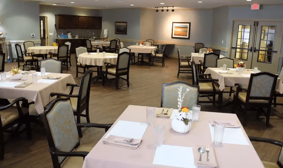 A dining room with multiple tables covered in light pink tablecloths, each set with placemats, utensils, glasses, and small floral centerpieces. The room has wooden flooring, beige walls, and a kitchenette area in the background. There are several chairs around each table and framed artwork on the walls.