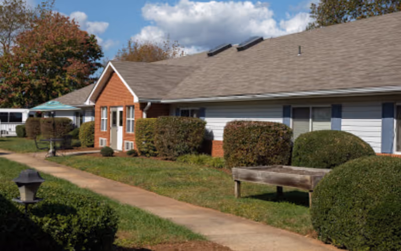 Exterior view of a single-story retirement facility building with a gray roof, white siding, and brick accents. The building is surrounded by neatly trimmed bushes and a green lawn. A concrete walkway runs alongside the building, and there is a wooden bench and a small outdoor table with an umbrella. Trees with autumn-colored leaves are visible in the background under a partly cloudy sky.