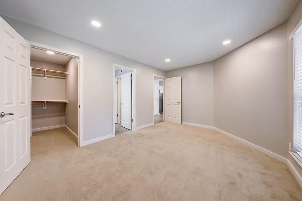 Empty carpeted bedroom with an open closet, two doors, recessed ceiling lights and a window.