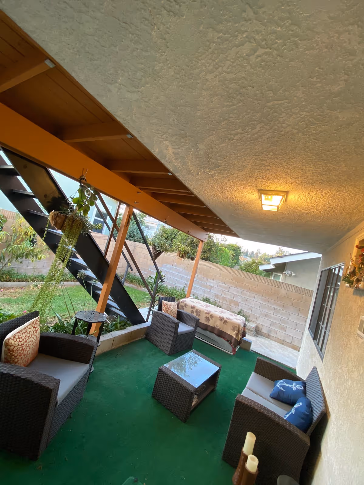 Covered outdoor patio area with green artificial grass flooring, featuring wicker furniture including two armchairs, a loveseat with blue pillows, and a glass-top coffee table. There is a hanging plant near a black metal staircase leading upstairs, and a bench covered with a floral blanket against a brick wall. The ceiling has a textured finish with a square light fixture.