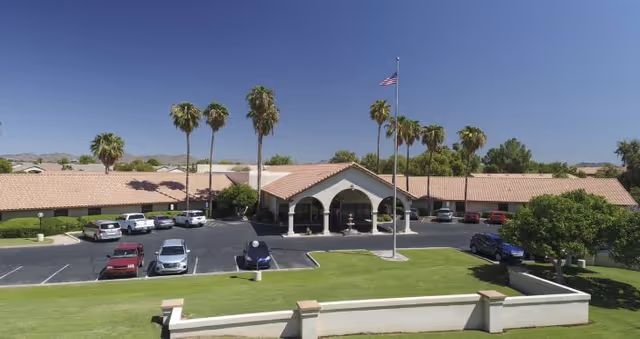Exterior view of Lake Pleasant Post Acute Rehabilitation Center showing a single-story building with a tiled roof, an arched entrance, a flagpole with an American flag, several parked cars, palm trees, and a well-maintained lawn under a clear blue sky.