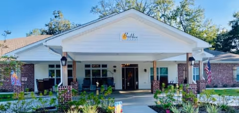 Front exterior view of BeeHive Homes Assisted Living building with a covered entrance, brick walls, white siding, and a garden with flowers and greenery in front. An American flag is displayed on the right side of the entrance.