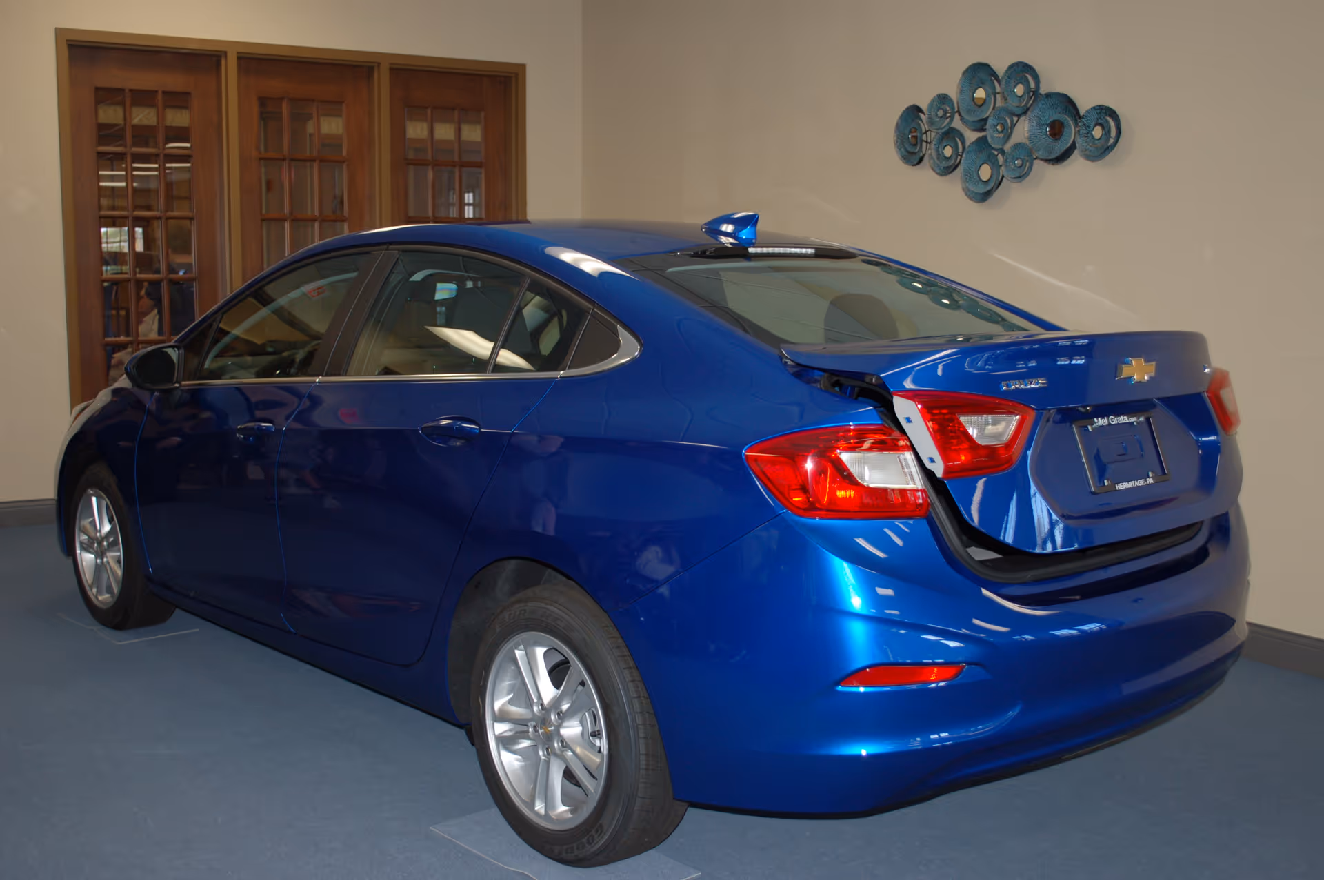A blue Chevrolet sedan parked indoors on a blue carpeted floor, with its trunk partially open. Behind the car are wooden double doors with glass panes and a wall decorated with a circular blue metal art piece.