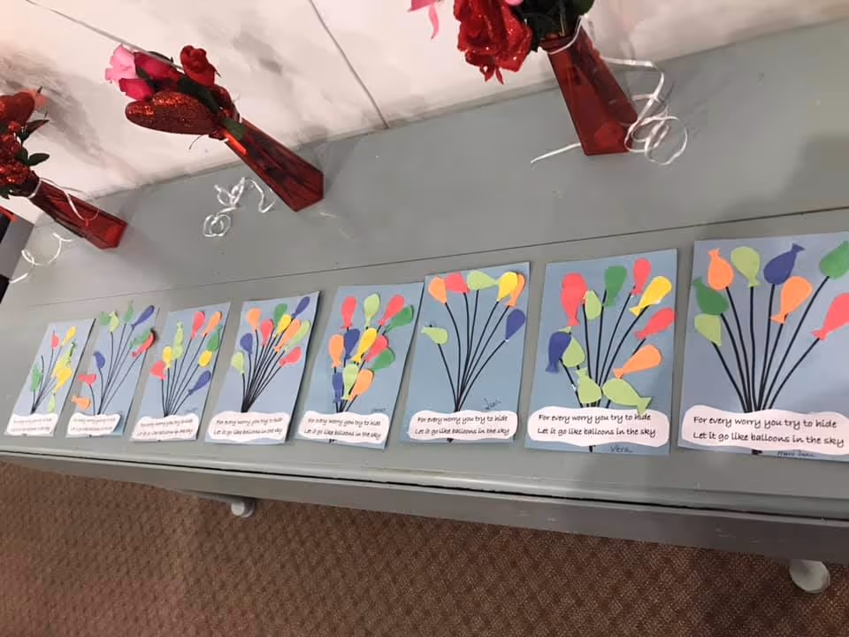 A row of handmade cards decorated with colorful paper balloons laid out on a table, with red vases of flowers behind them.