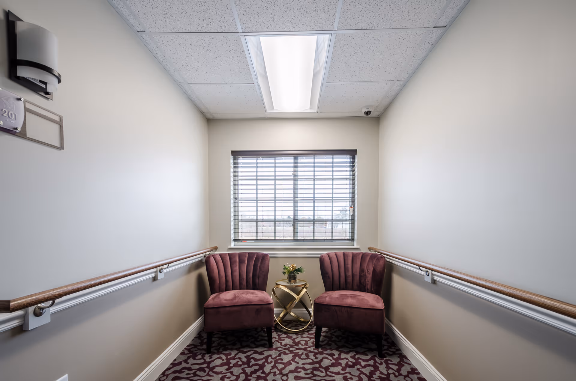 A small seating area in a hallway with two purple upholstered chairs and a small round gold table with a flower arrangement between them. The walls are beige with wooden handrails on both sides, a window with blinds at the end, and a ceiling light above.