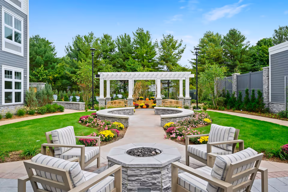 Outdoor seating area with four cushioned chairs arranged around a stone fire pit, surrounded by green grass, colorful flowers, and a paved walkway leading to a white pergola with benches underneath. Trees and a gray fence border the area.