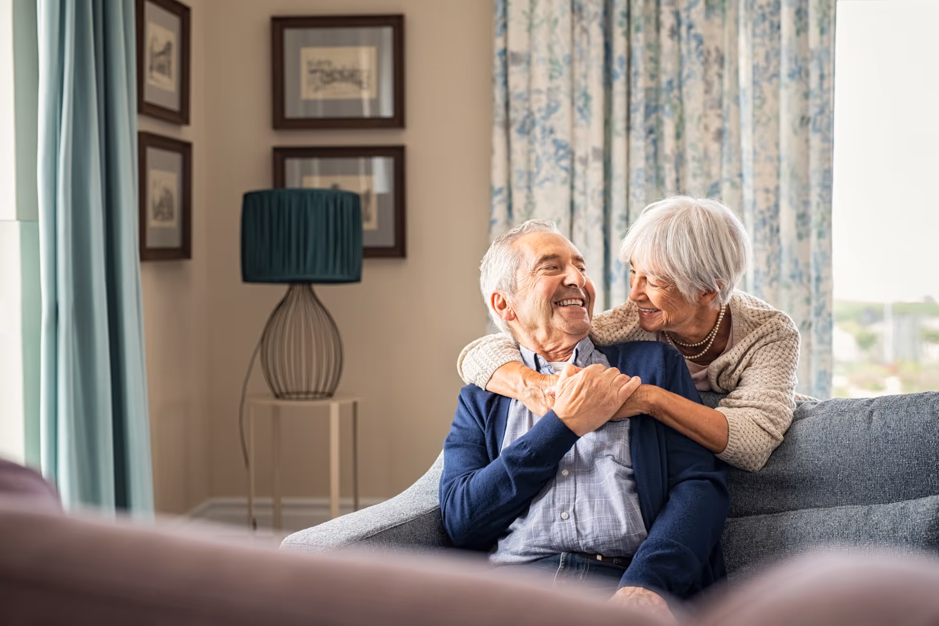 An elderly couple sitting on a gray sofa in a cozy living room. The woman is hugging the man from behind, and both are smiling warmly at each other. The room has light-colored walls, framed pictures, a modern lamp, and floral curtains.