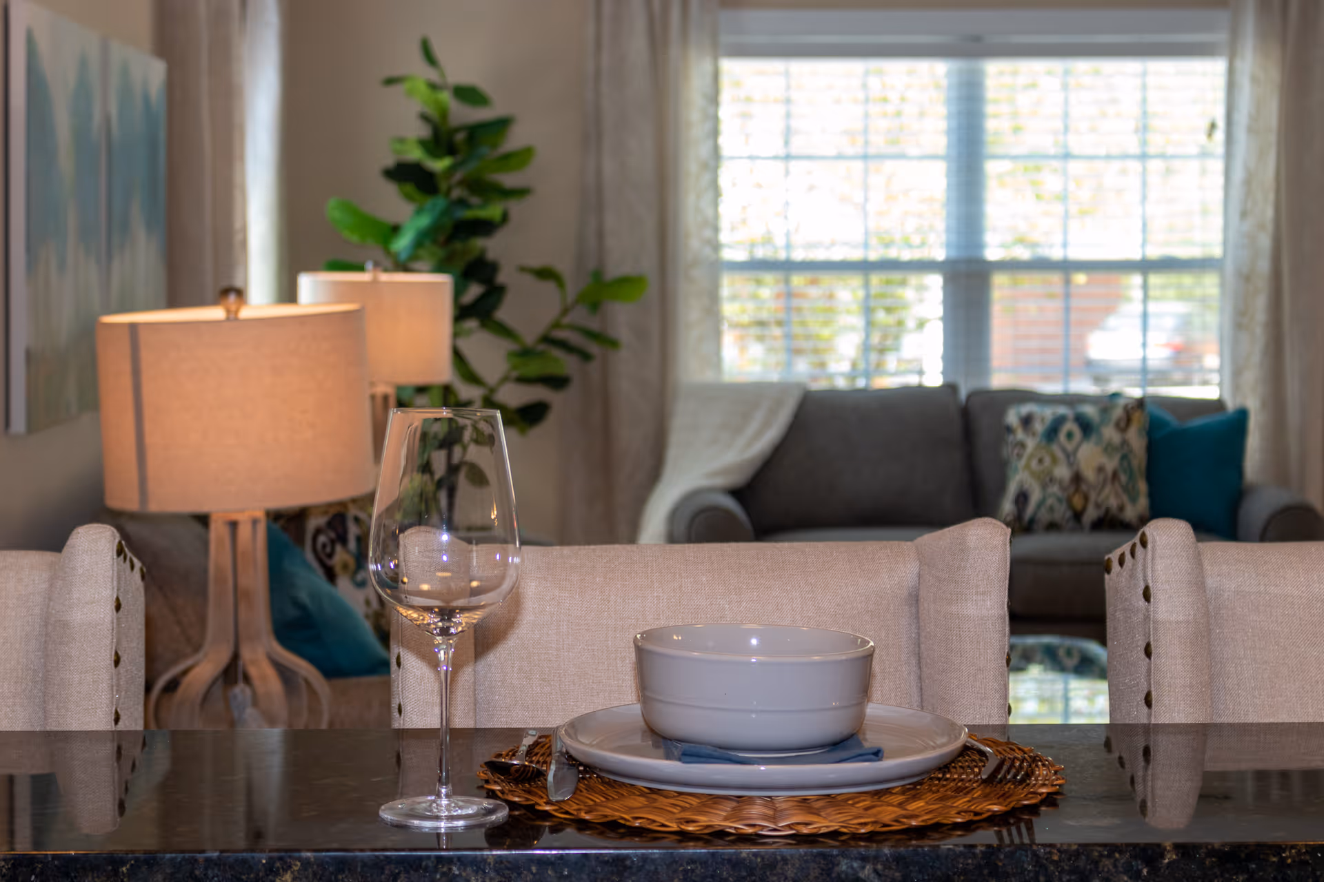 A cozy living room and dining area with a set table featuring a white bowl on a plate with a woven placemat and a wine glass. In the background, there is a gray sofa with patterned and solid pillows, a floor lamp, a green leafy plant, and a large window with blinds letting in natural light.