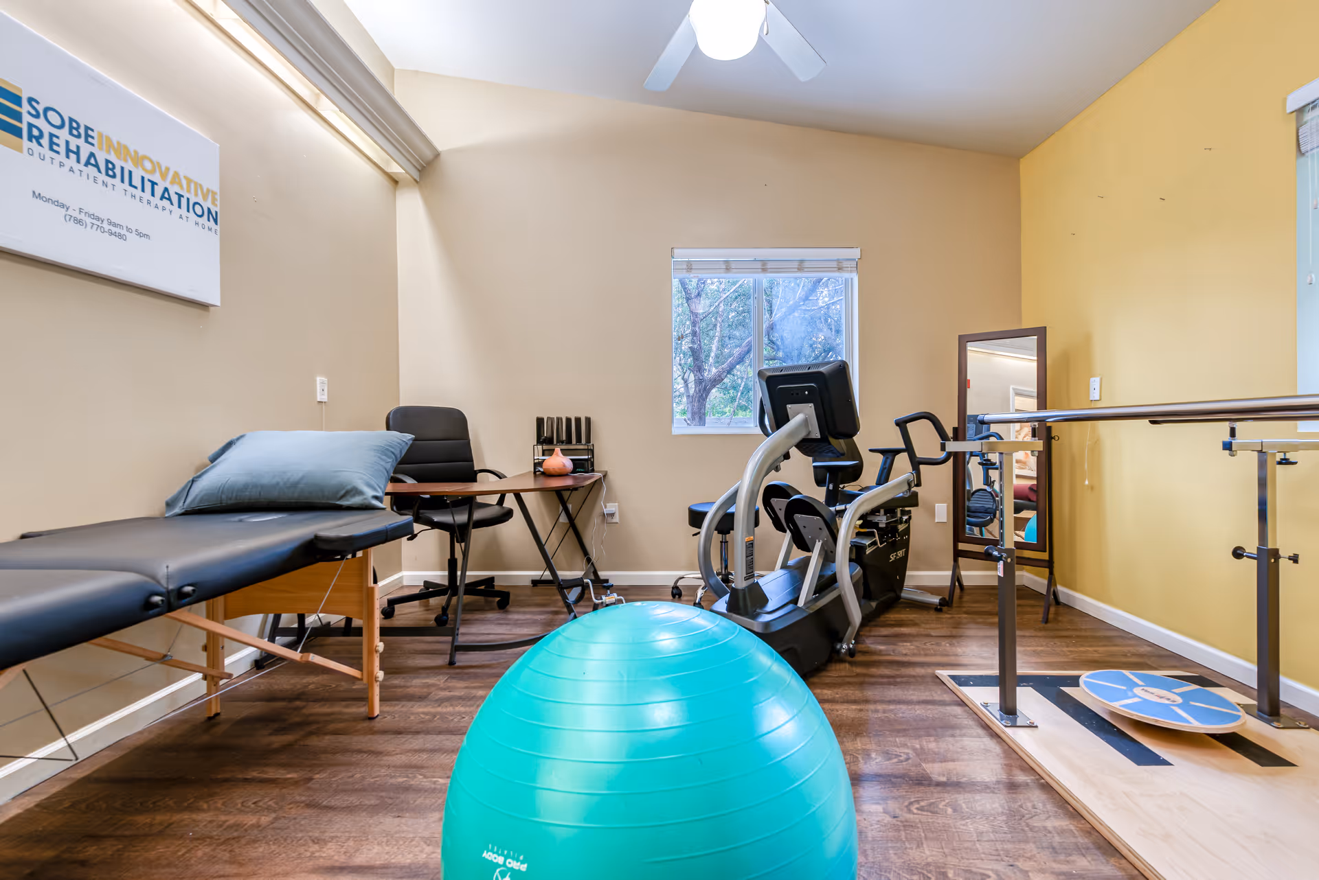 Physical therapy room with a treatment table, office chair and desk, exercise bike, balance board with parallel bars, large green exercise ball, and a window showing trees outside. A sign on the wall reads 'Sobe Innovative Rehabilitation Outpatient Therapy at Home'.