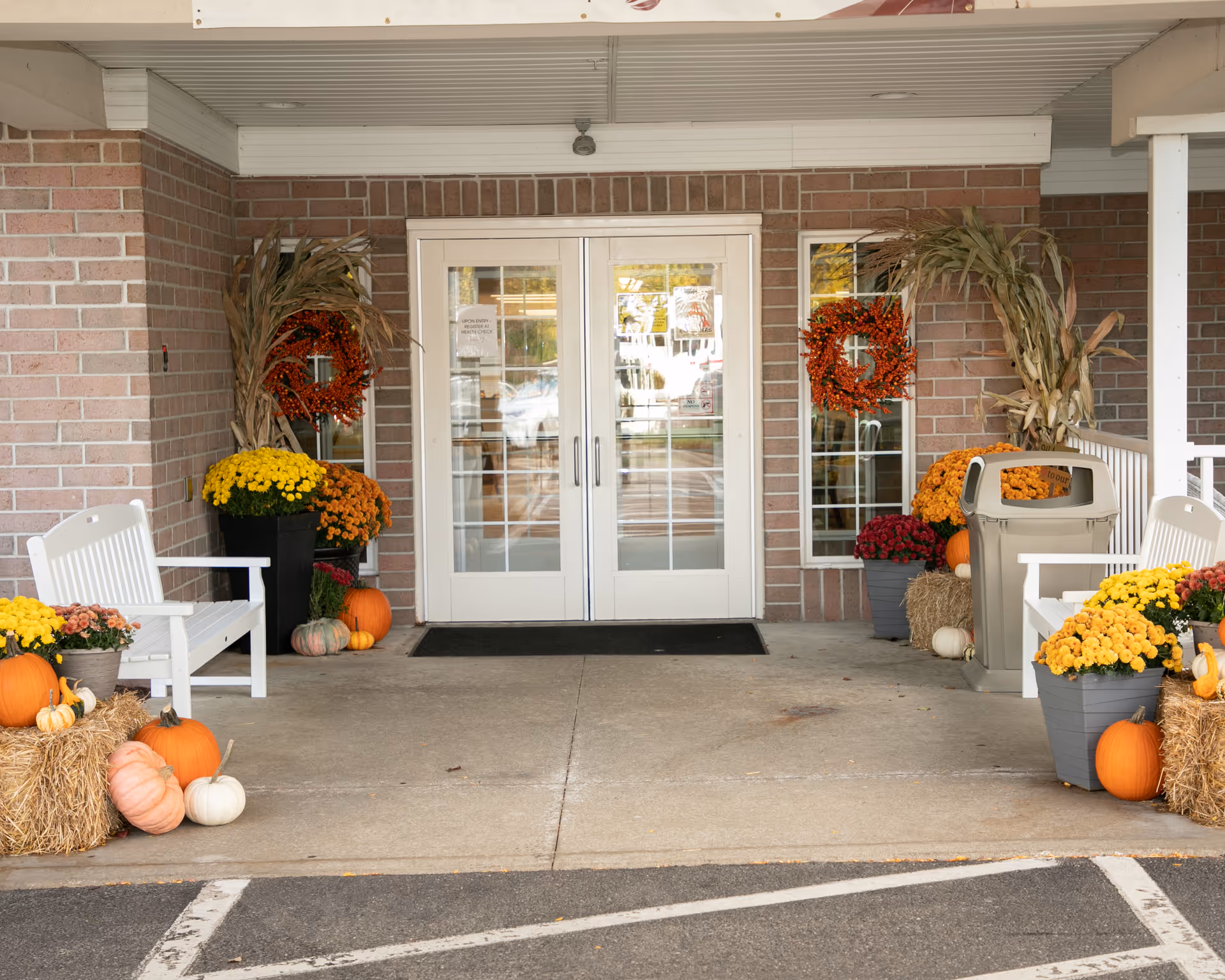 Entrance to a building with double glass doors, decorated with autumn-themed items including pumpkins, hay bales, yellow and orange flowers, corn stalks, and orange wreaths on the windows. White benches are placed on either side of the entrance, and a trash bin is visible on the right side.