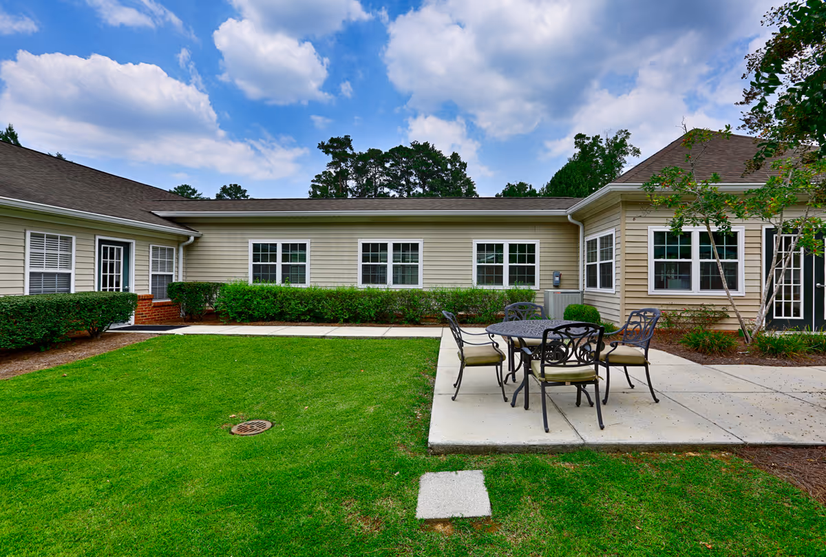 A green courtyard with a round metal patio table and chairs on a concrete slab in front of a single-story beige building under a partly cloudy sky.