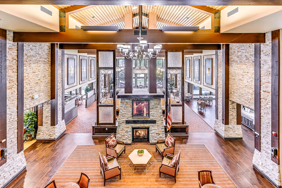 Spacious senior living facility common area with a high wooden ceiling, large stone fireplace in the center, surrounded by four cushioned chairs with patterned pillows and a small hexagonal table. The room features wood flooring, stone accent walls, large windows, and a chandelier hanging from the ceiling. Adjacent dining areas with tables and chairs are visible through open doorways on either side.