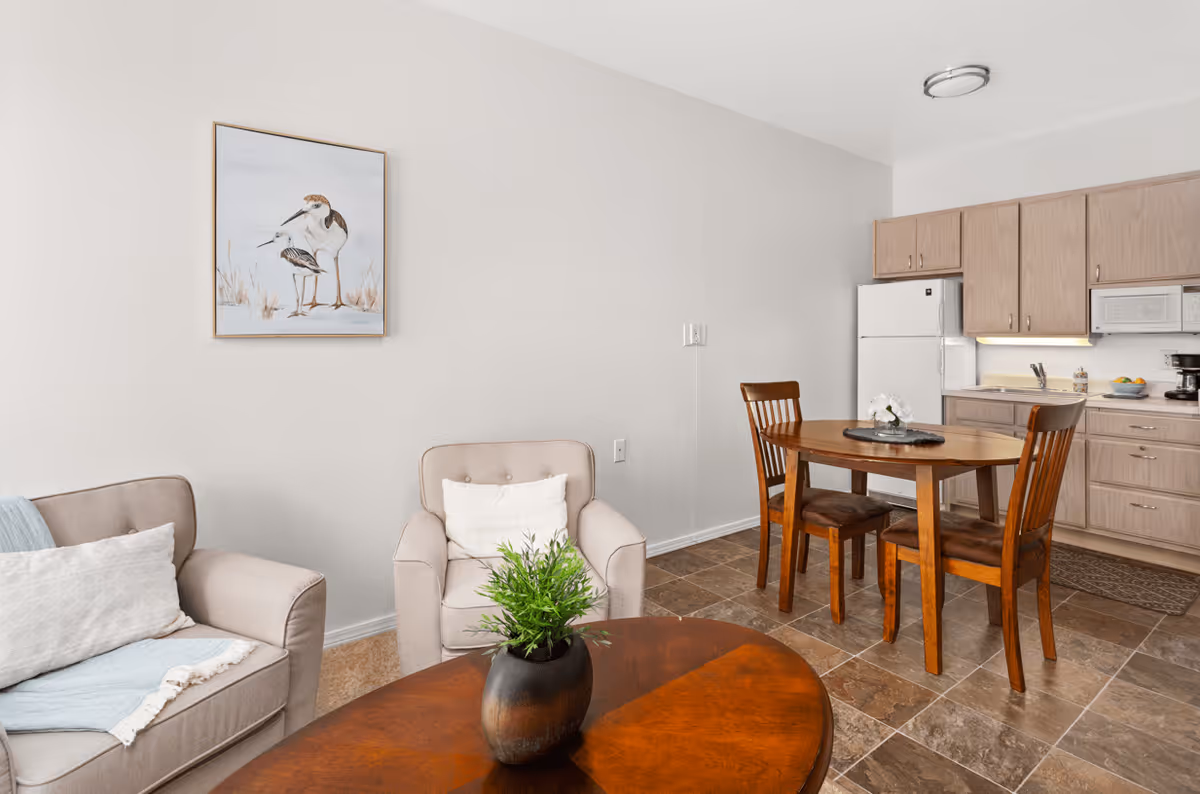 A cozy living and dining area with two beige armchairs, a wooden coffee table with a potted plant, a wooden dining table with three chairs, and a kitchen area with light wood cabinets, a white refrigerator, microwave, and coffee maker. A framed picture of birds hangs on the wall.