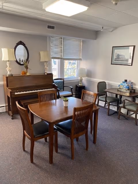 A cozy room with a wooden piano against the wall, flanked by two table lamps and a small oval mirror above it. In the center, there is a wooden table with four chairs around it, and a small potted plant on the table. Near the window, there are two chairs and a small table with board games stacked on it. The room has carpeted flooring, white walls with a chair rail, and a framed picture hanging on the wall.