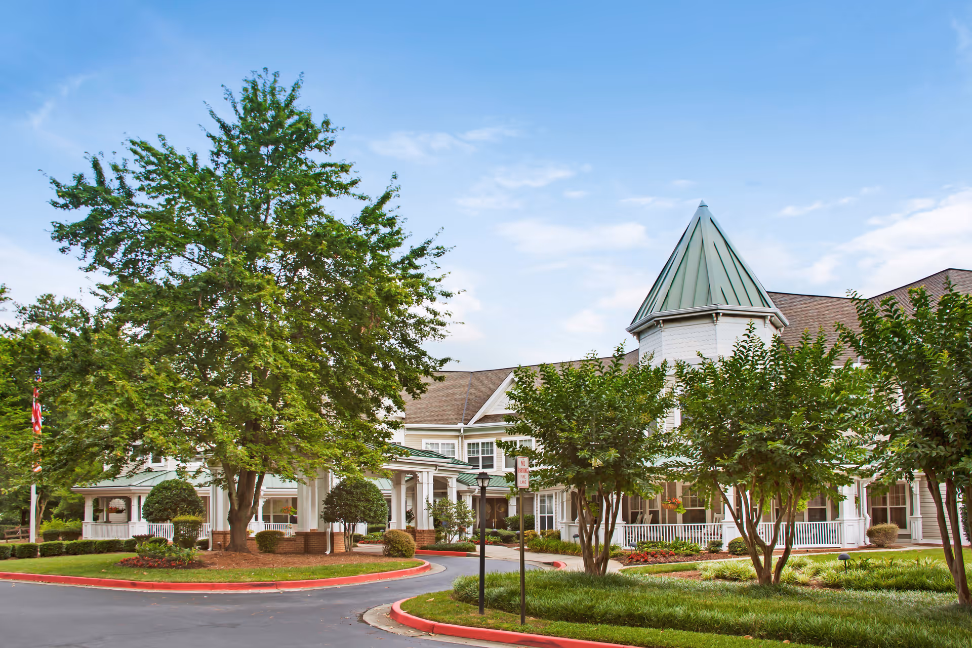 Exterior view of a senior living facility with a circular driveway, well-maintained landscaping including trees and shrubs, and a building with a green pointed roof tower under a blue sky.