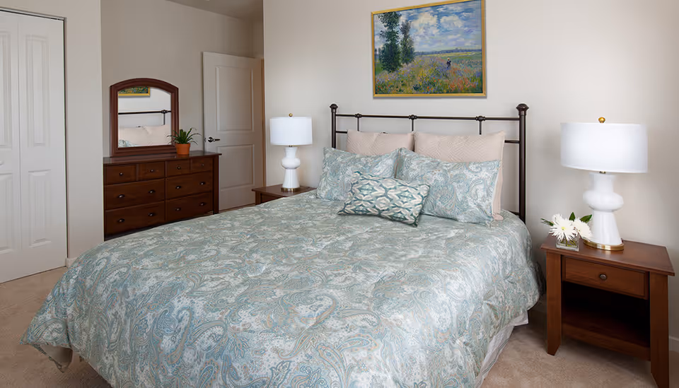 A neatly made bed with a blue and beige paisley patterned comforter and matching pillows in a bedroom. On each side of the bed are wooden nightstands with white lamps, and one nightstand has a small vase with white flowers. A wooden dresser with a mirror and a small potted plant is visible against the wall. A framed painting hangs above the bed, and there are two closed doors in the background.