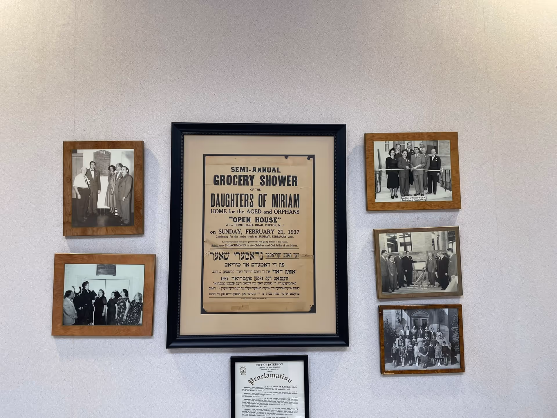 A wall display featuring six framed black and white photographs and two framed documents. The central large document is a vintage poster for a Semi-Annual Grocery Shower event for the Daughters of Miriam Home for the Aged and Orphans dated February 21, 1937. Surrounding it are various historical photos of groups of people, likely related to the facility's history.
