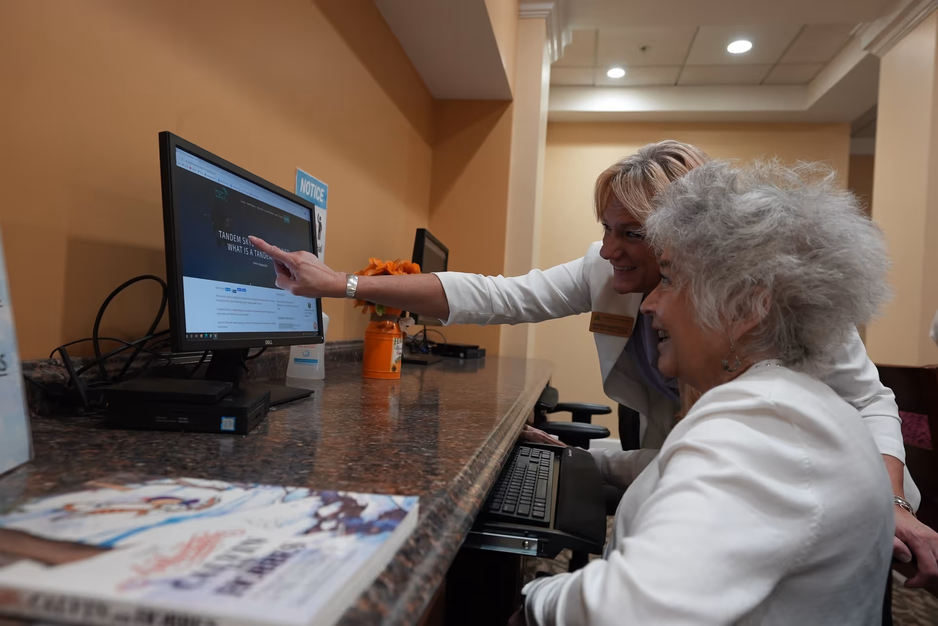 Two women, one older and one younger, are sitting at a computer workstation in an indoor setting. The younger woman is pointing at the computer screen while the older woman looks on smiling. The workspace has a granite countertop with a keyboard tray, and there are some books and a small orange flower pot on the counter.