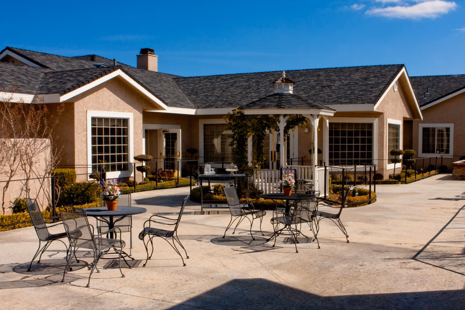 Outdoor patio area at Sterling Inn Assisted Living Community featuring metal tables and chairs with potted flowers, a white gazebo with greenery, and beige buildings with large windows under a clear blue sky.