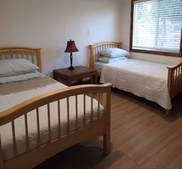 A simple bedroom with two single wooden beds, each made with white bedding and pillows. Between the beds is a wooden nightstand with a red lampshade lamp. A window with white blinds is on the right wall, allowing natural light into the room. The floor is wooden.