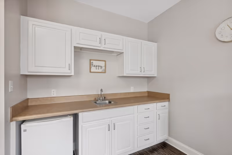 A small kitchenette area with white cabinets, a beige countertop, a small sink, and a mini refrigerator underneath the counter. A decorative sign that says 'happy' is mounted on the wall above the sink. A round clock is partially visible on the right wall.