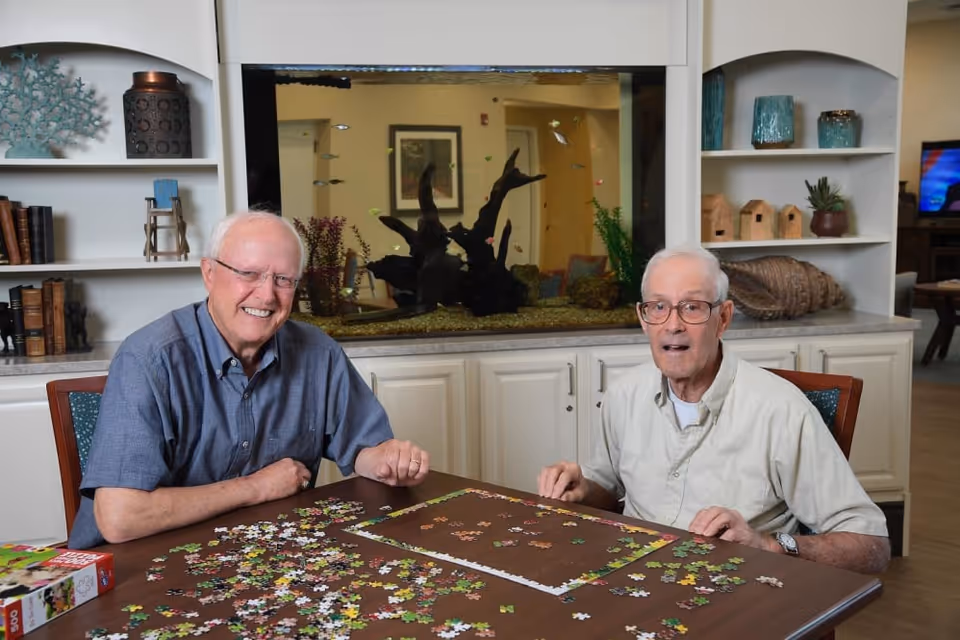 Two elderly men sitting at a table working on a jigsaw puzzle together in a well-lit room with a large fish tank and shelves with decorative items in the background.