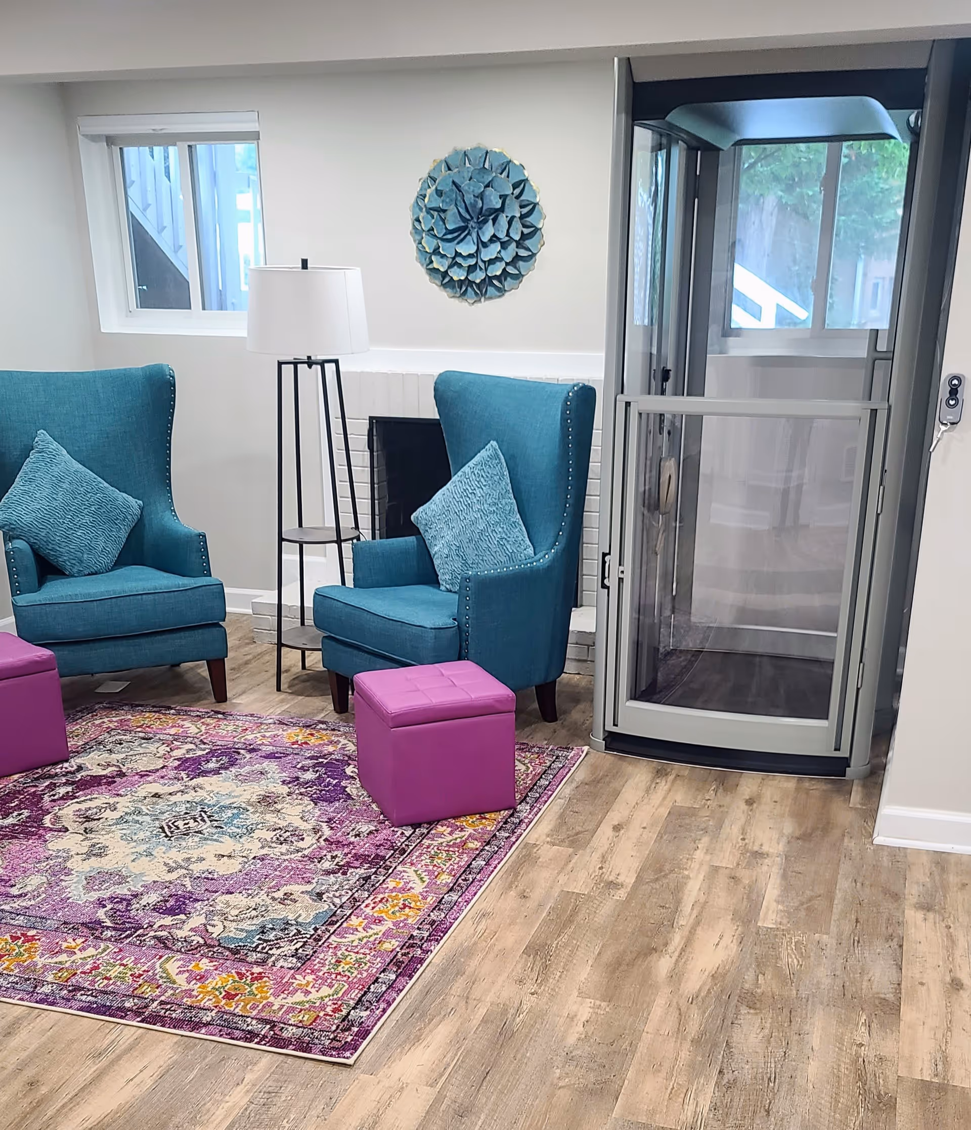 A cozy living room area with two teal armchairs, each with a matching cushion, a colorful purple and cream patterned rug, two small purple ottomans, a tall floor lamp with a white shade, a decorative blue wall art piece, and a glass enclosed elevator or lift next to a window.