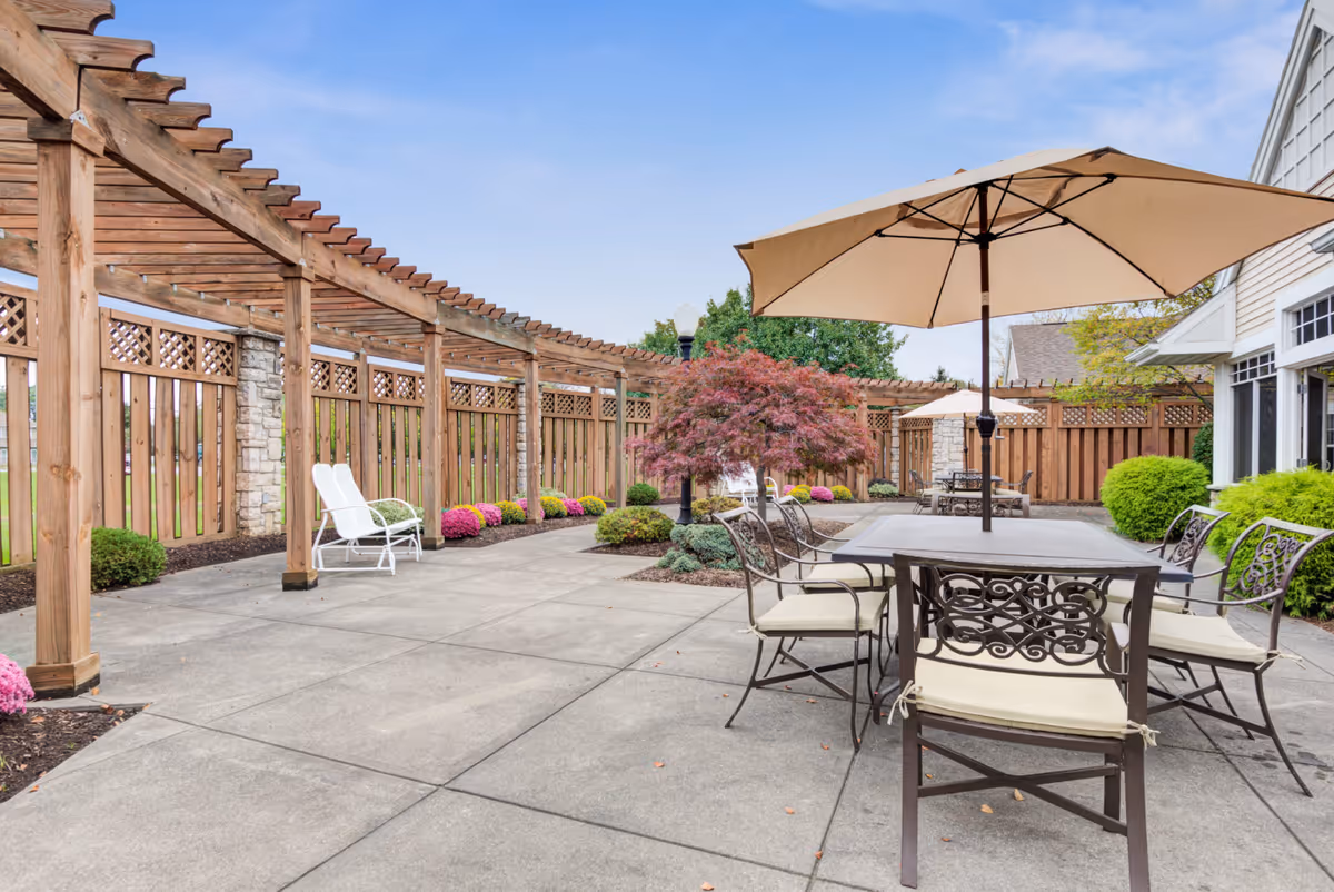 Outdoor patio area with a large table and six chairs under a beige umbrella. There is a wooden pergola along the left side with two white lounge chairs underneath. The patio is surrounded by a wooden fence with stone pillars and landscaped with bushes and colorful flowers. A small tree with red leaves is near the center of the image.